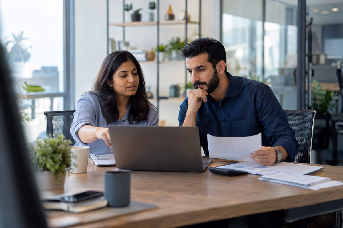 Two finance professionals reviewing accounting records and a laptop at a bright office table